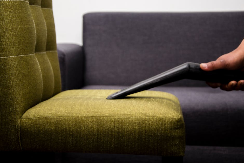 Close-up of a person using a handheld vacuum cleaner to clean a green upholstered armchair, with the vacuum nozzle pressed into the fabric. In the background, a dark grey sofa is visible in a well-lit