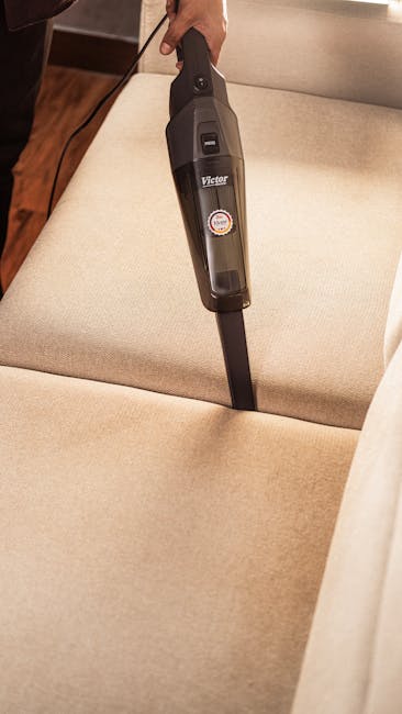 Close-up of a person using a handheld vacuum cleaner to clean a green upholstered armchair, with the vacuum nozzle pressed into the fabric. In the background, a dark grey sofa is visible in a well-lit